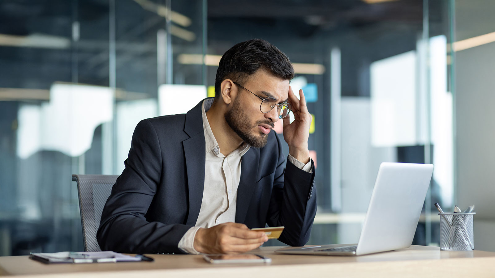 A businessman expresses stress as he holds a credit card while working on a laptop