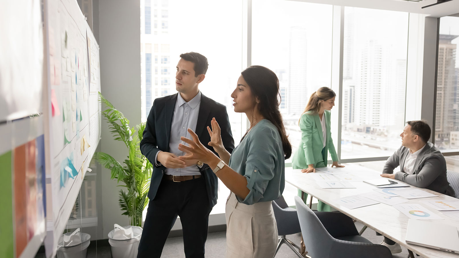In modern office with city view, woman and man standing near whiteboard filled with sticky notes