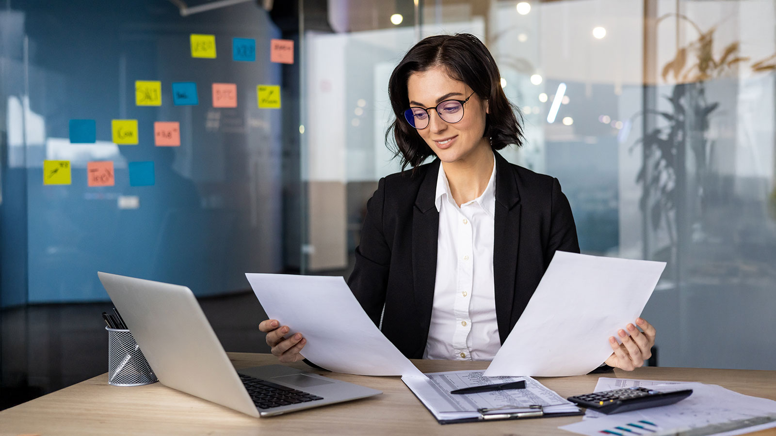Professional businesswoman reviewing documents at desk with laptop in modern office space