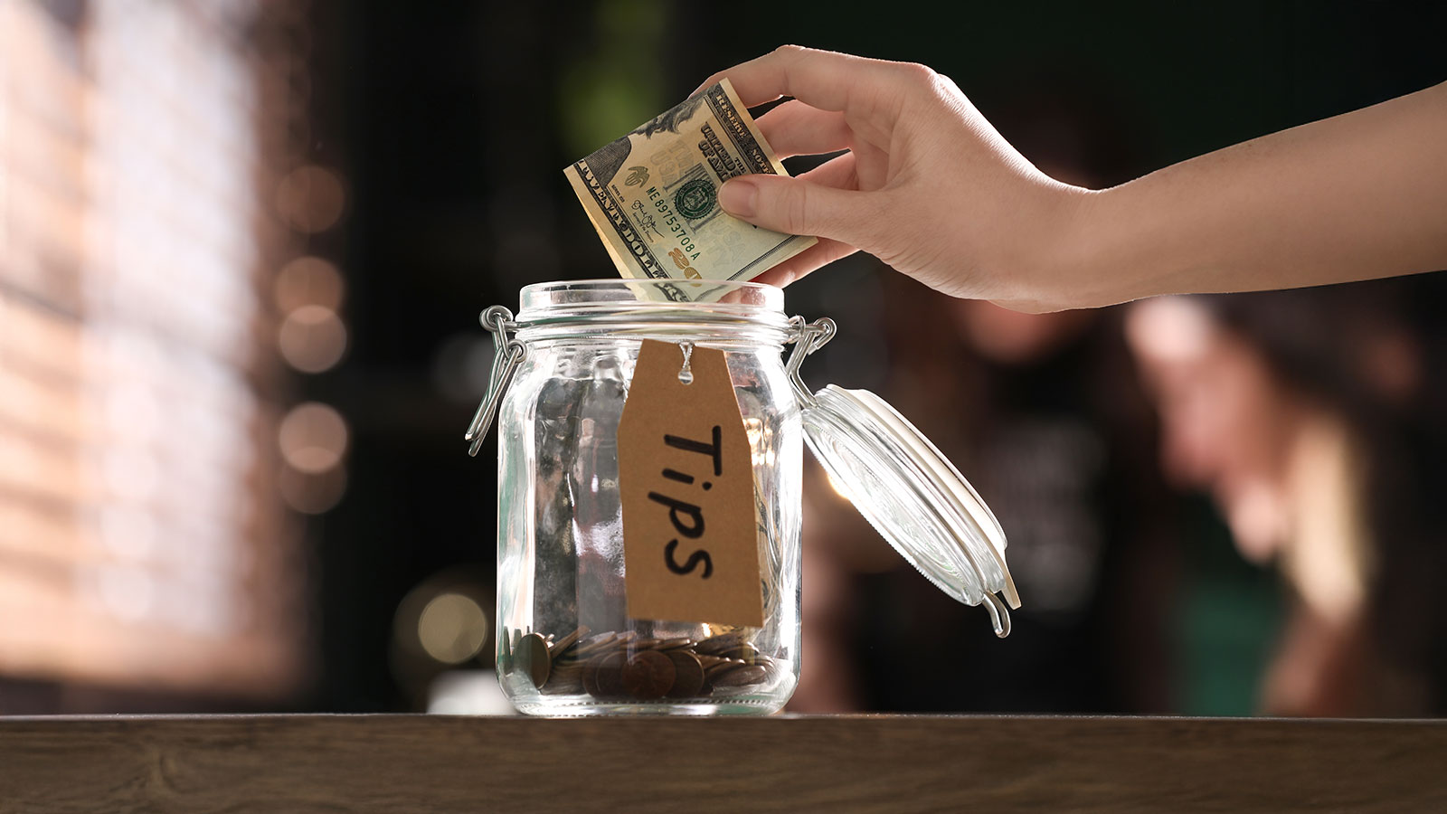 Woman putting tips into glass jar on wooden table indoors, closeup