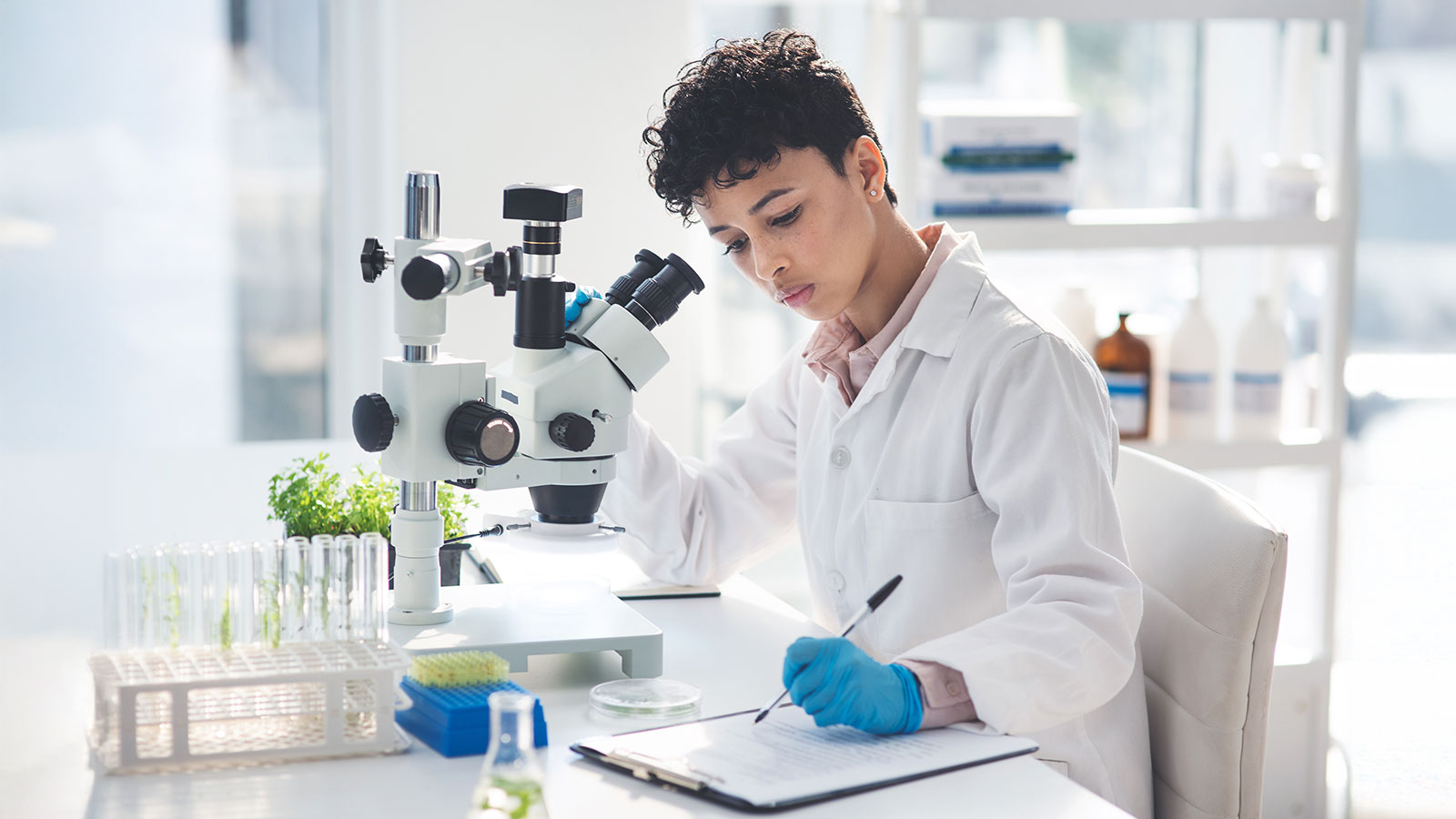 young female scientist making notes while working with a microscope and plants in a laboratory