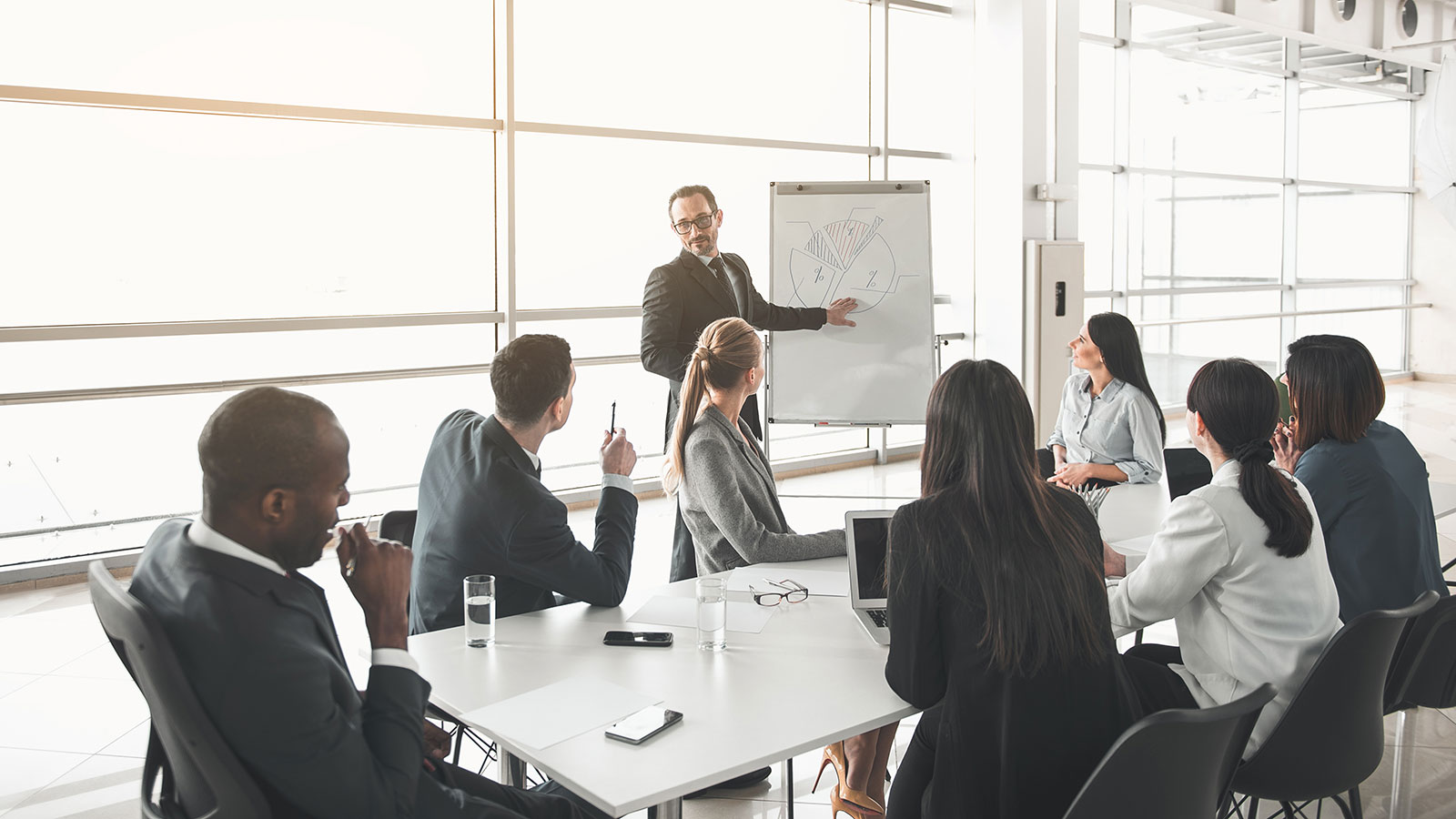 Young man standing in front of audience near marker board and pointing at chart written on it